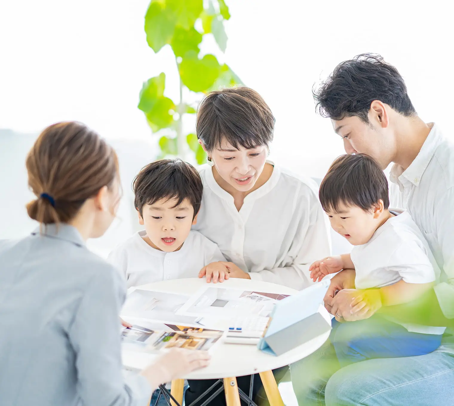 明るい室内で、4人家族(父親、母親、2人の子供)が女性スタッフと資料を囲んで相談している様子。テーブル上には住宅関連の資料やカタログが広げられ、観葉植物も見える。マイホーム購入や不動産の相談風景。